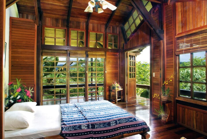 A serene wooden bedroom with large windows, a double bed featuring a blue patterned blanket, and lush greenery visible outside. The room has a high ceiling with a ceiling fan and several potted plants—a perfect retreat for those seeking tranquility in Costa Rica.