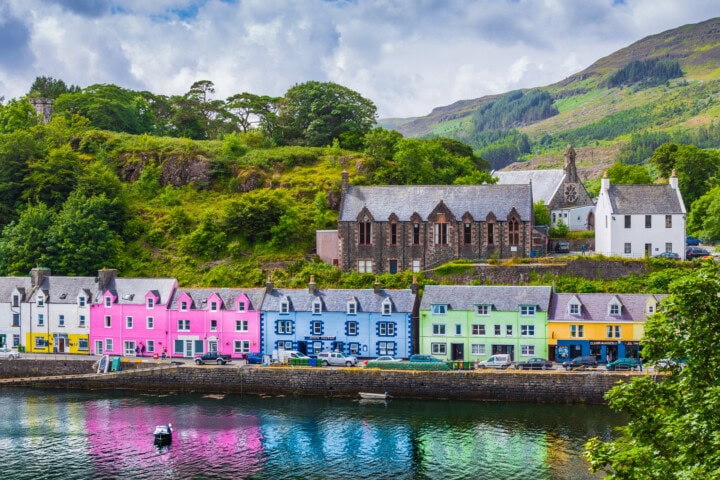 A row of colorful houses along a waterfront harbor in Scotland, with green hills, trees, and a church in the background. The buildings reflect in the calm water below, creating a picturesque scene perfect for travel and tourism.