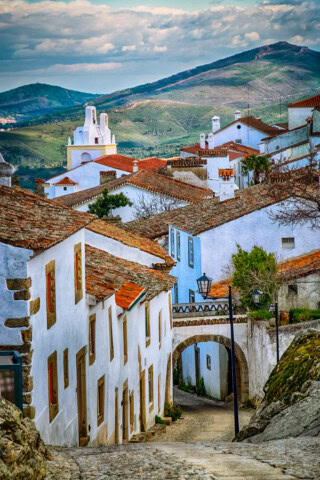 Narrow cobblestone street lined with white buildings featuring red-tiled roofs. Archway and lampposts are visible, reminiscent of Portugal, with green hills and mountains in the background under a partly cloudy sky.