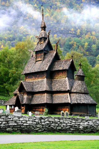 A wooden stave church surrounded by trees and mist, with a stone wall and gravestones in the foreground, stands as a testament to Norway’s architectural heritage, an enchanting destination for travel and tourism enthusiasts.