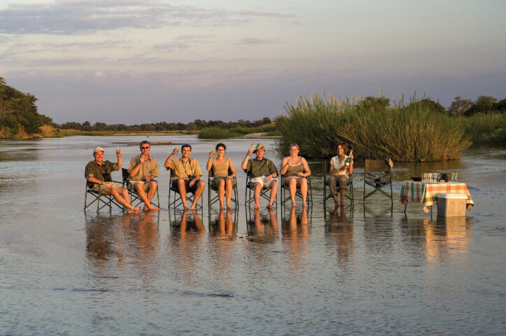A group of six people sits on folding chairs in shallow water, surrounded by Zambia's natural scenery, with a table to the right holding items and a checked cloth.