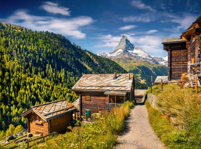 Wooden houses on a pathway with lush green hills and the Matterhorn mountain in the background, all under a partly cloudy sky, showcase the quintessential charm of Switzerland's tourism landscape.