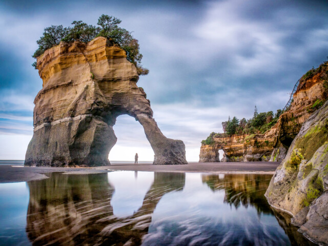 A person stands near a large rock formation with an arch at a beach in New Zealand. The overcast sky is reflected in the shallow water, while vegetation grows atop the rock.