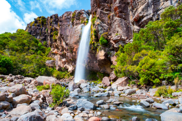 A waterfall cascades down a rocky cliff into a stream surrounded by lush green foliage and boulders under a partly cloudy New Zealand sky.