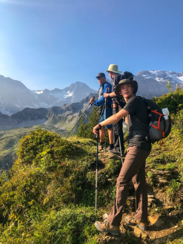 Three hikers, equipped with backpacks and trekking poles, stand on a mountain trail in Switzerland, gazing at the scenic view of distant snow-capped peaks under a clear blue sky.