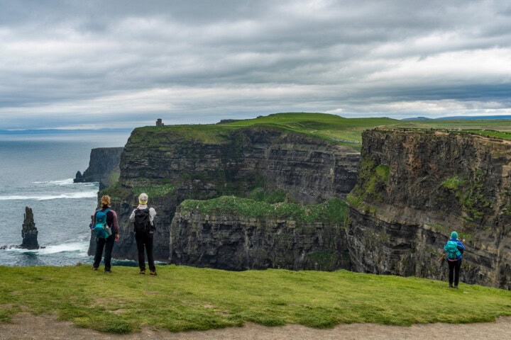 Three people stand on a grassy cliff in Ireland, overlooking the Cliffs of Moher. The ocean and dramatic rocky formations stretch out under a cloudy sky.