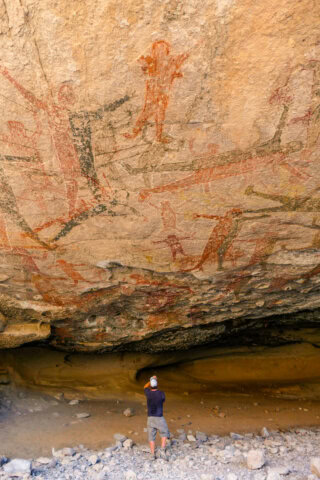 A person stands beneath a rock face in Mexico, adorned with ancient cave paintings, appearing to take a photo of the artwork.