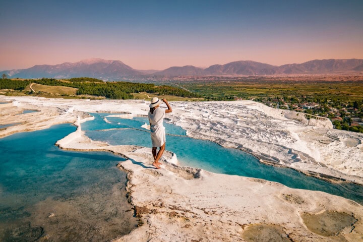 A person in a white dress and hat stands at the edge of a turquoise mineral pool on a white, rocky landscape in Turkey, with mountains and a valley in the background under a clear sky, creating an idyllic scene perfect for travel enthusiasts.