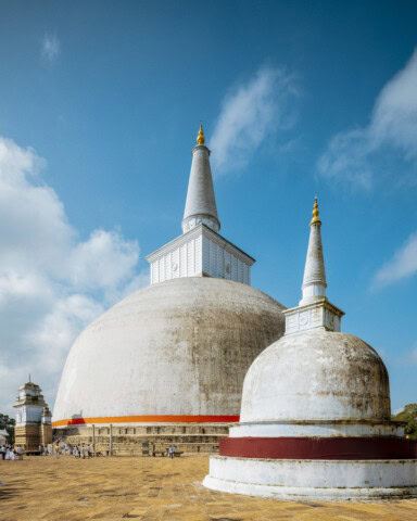 Large white stupa with a smaller stupa in front, set against a blue sky with scattered clouds; people at the base for scale.