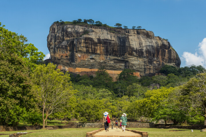Three people walk on a path towards a large rock formation surrounded by trees at Sigiriya, Sri Lanka, on a clear day.