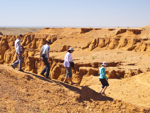 Four people walking along a ridge in a desert landscape with eroded rock formations in the background, reminiscent of Mongolia's vast and ancient terrains.