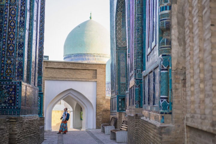 Woman walking through a narrow alley with intricate blue and green tilework, leading to a domed structure in the background.