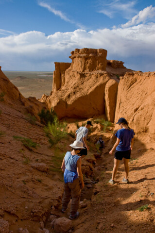 Four people hike down a rugged, reddish-brown canyon under a blue sky with clouds, reminiscent of Mongolia's vast landscapes.