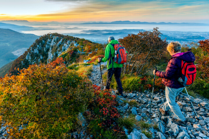 Two hikers with backpacks and trekking poles traverse a rocky mountain trail surrounded by autumn foliage during sunrise, with a misty valley and distant mountains in the background, showcasing the serene beauty of Slovenia—a perfect destination for nature enthusiasts and travel aficionados.