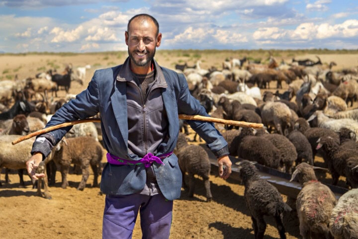 A man stands smiling in front of a flock of sheep in an Uzbekistan desert landscape, holding a wooden staff across his shoulders. He wears a jacket, a purple belt, and has a shaved head.