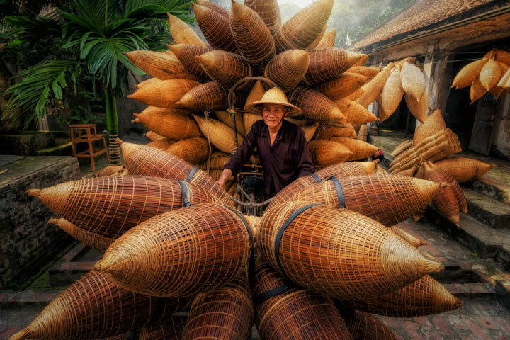 A person wearing a conical hat stands behind a bicycle loaded with numerous woven baskets in an outdoor setting, capturing the essence of daily life in Vietnam.