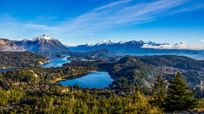A scenic mountain landscape in Argentina with snow-capped peaks, multiple lakes, and lush green forests under a clear blue sky.