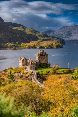 A historic castle stands on a small island in Scotland, connected by a bridge, surrounded by water and lush, green hills under a cloudy sky.