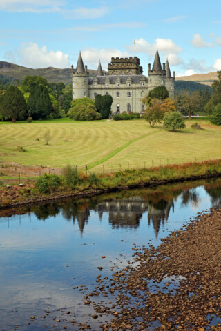 A large historic castle with multiple towers and turrets sits on a grassy hill in Scotland, bordered by trees and reflected in a calm river in front.