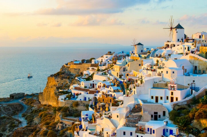 A coastal village in Greece with white buildings and windmills overlooks the sea at sunset, with a sailboat visible in the distance.