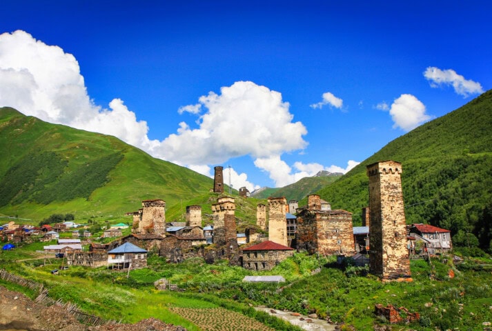 A scenic view of a historic Georgian village with stone towers and houses set against a backdrop of green mountains and blue sky with clouds.