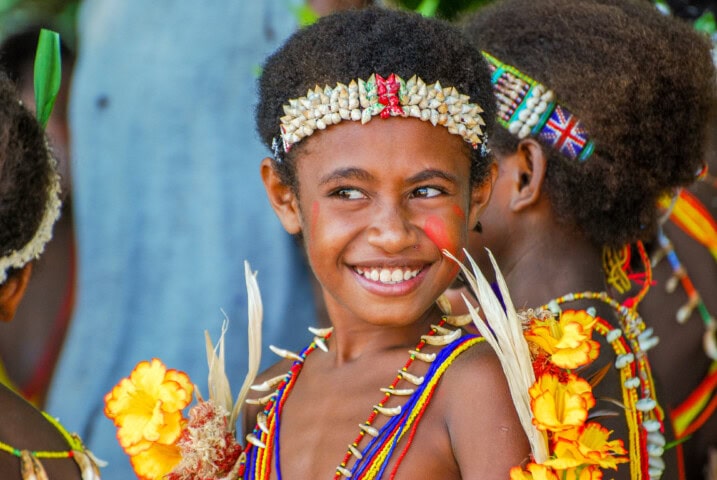 Young child in traditional Papua New Guinea attire with a beaded headdress, face paint, and colorful decorations, smiling and standing among others wearing similar outfits.