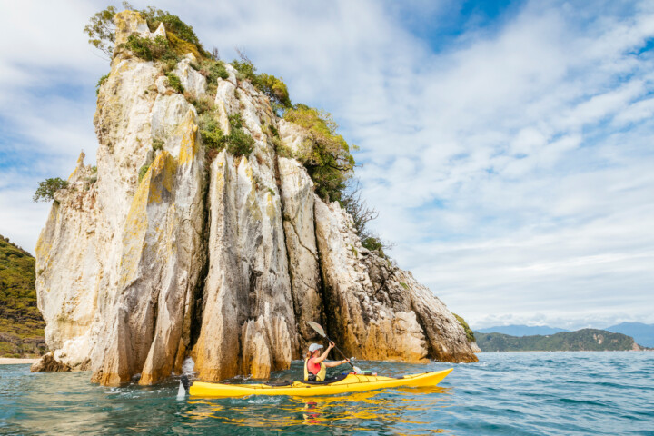 A person in a yellow kayak paddles near a large, rocky outcrop in the clear blue waters of New Zealand, under a partly cloudy sky.
