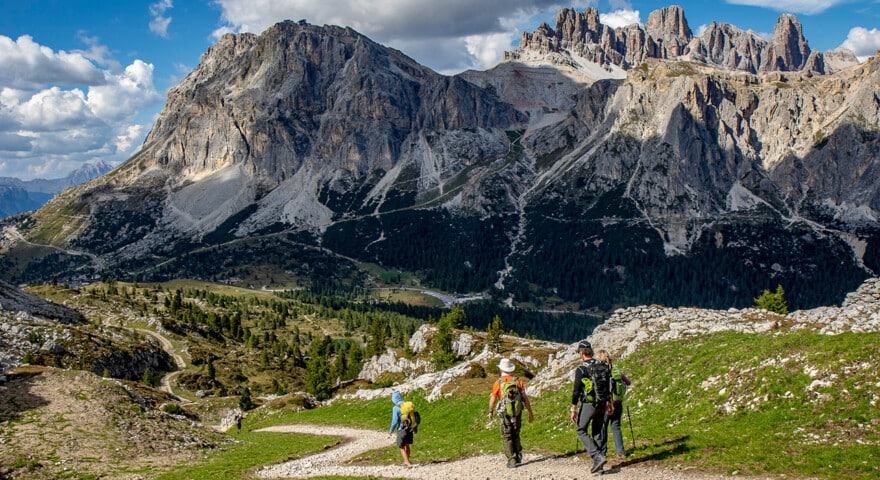 Four hikers walk on a trail through the mountainous landscape of the Dolomites, with rocky peaks and grassy areas under a partly cloudy sky—a perfect scene for travel enthusiasts and tourism admirers alike.