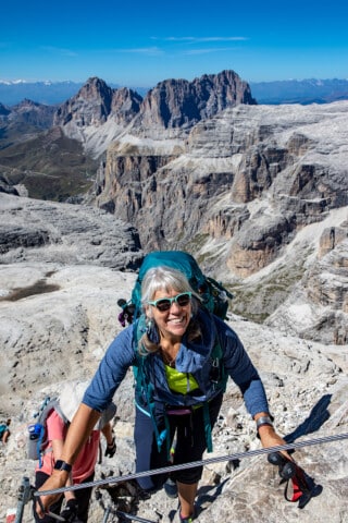 A person climbs a rocky mountain path equipped with a backpack; visible in the background are the rugged peaks of the Dolomites and a clear blue sky.