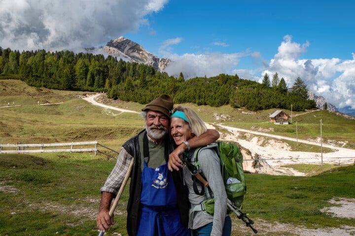 Two elderly hikers share an embrace on a lush mountain trail in the Italian Alps, with a cabin and forested hills in the background under a partly cloudy sky.