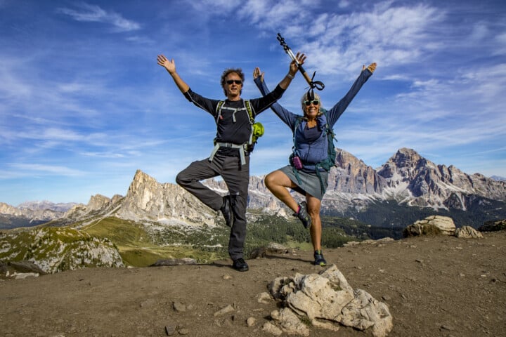 Two hikers pose in a tree yoga pose on a rocky mountain trail in the Dolomites, their hiking gear and the stunning Italian mountain landscape framed by a blue sky with scattered clouds.