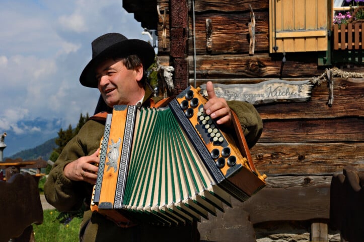 A person wearing a hat and traditional attire plays an accordion in front of a rustic wooden building with a small window and a sign, nestled in the scenic mountains of the Dolomites, Italy.