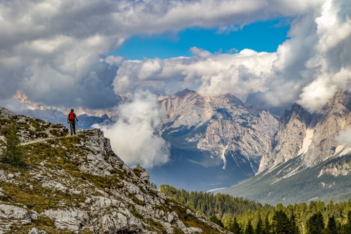 A person wearing a red jacket stands on a rocky cliff overlooking the vast, cloud-covered peaks of the Dolomites.