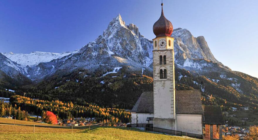 A picturesque church with a red dome stands in a grassy field, set against the backdrop of the majestic Dolomites and a small Italian town nestled at their base.