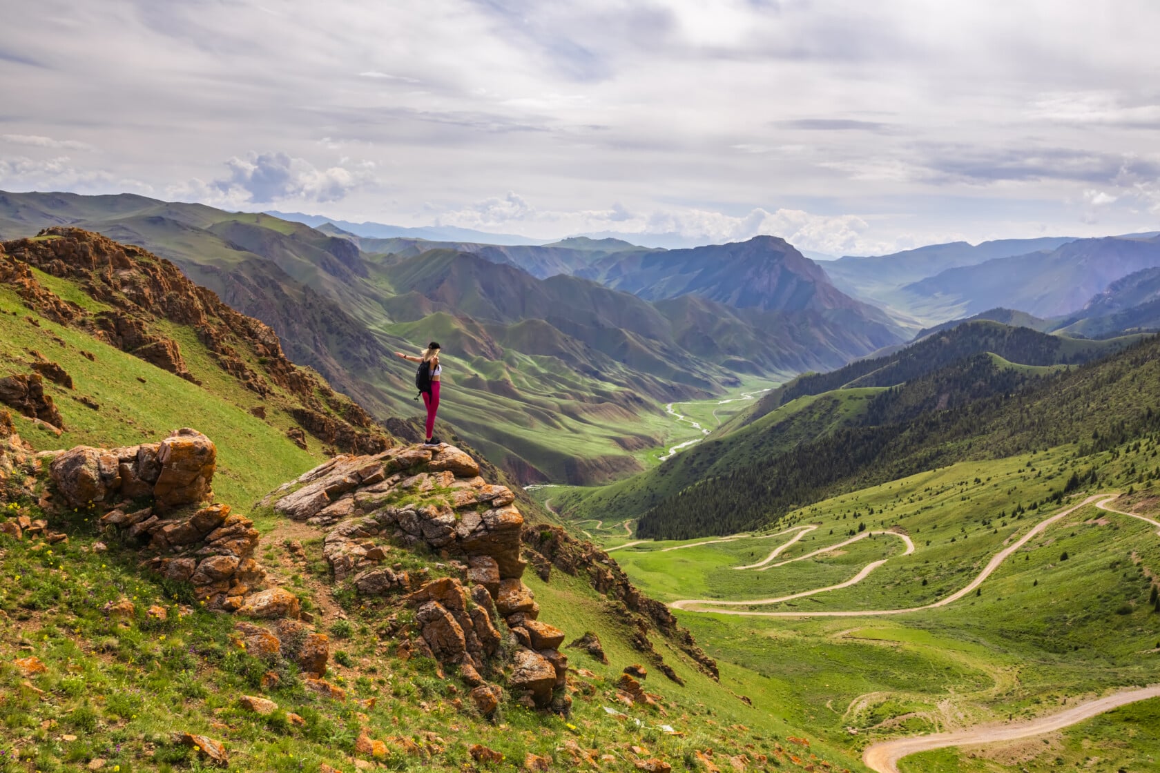 A person stands on a rocky outcrop overlooking a vast, green mountainous landscape with winding paths and a river in the valley below under a partially cloudy sky, pondering the best time to visit this breathtaking scenery.