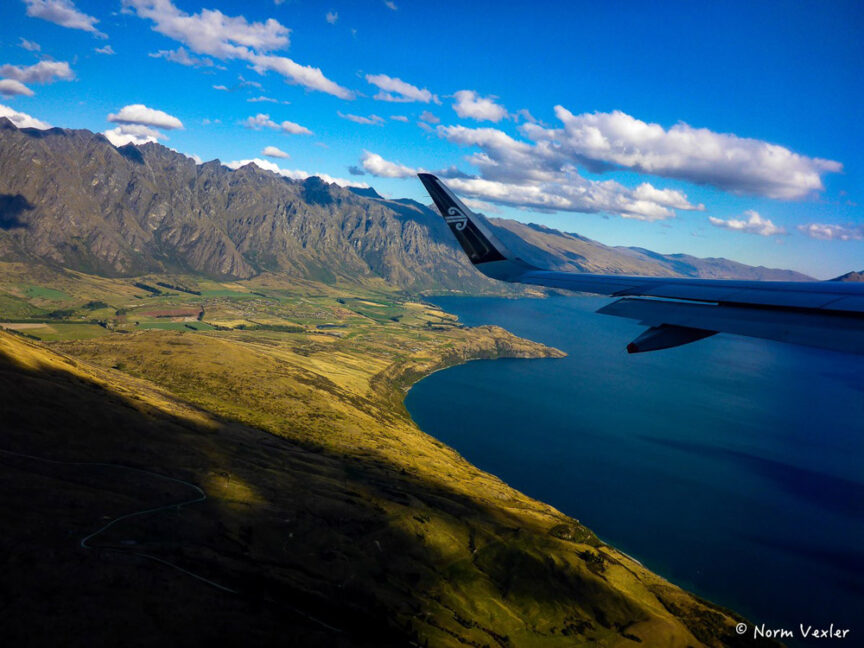 Aerial view from an airplane window showing a wing, rugged mountains, and a large body of water with coastline under a blue sky with scattered clouds, resembling the varied landscapes of Middle Earth.