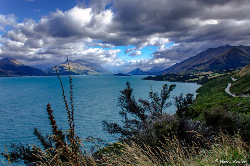 A landscape reminiscent of Middle Earth features a blue lake surrounded by mountains under a partly cloudy sky, with shrubs and tall grasses in the foreground along a winding coastline.