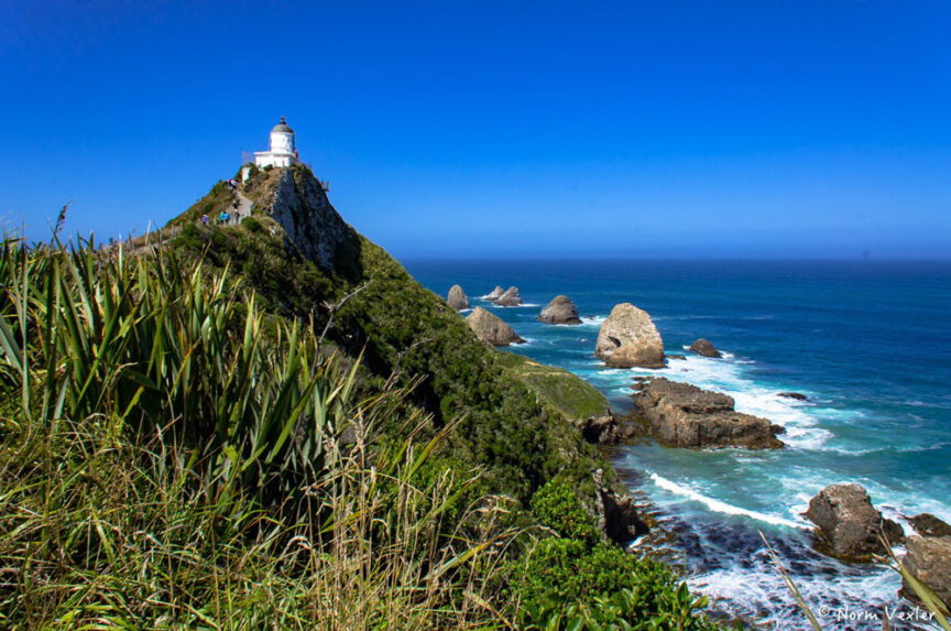 A lighthouse sits atop a rocky hill, much like those found in Middle Earth, overlooking the ocean with waves crashing against the rocks below and vegetation in the foreground under a clear blue sky.