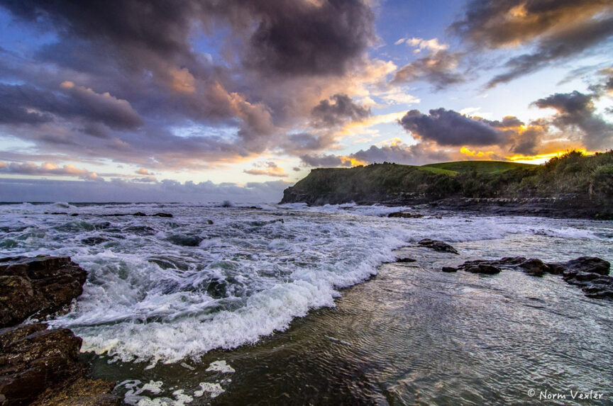 A coastal landscape at sunset features waves crashing against rocky shorelines, a distant hill reminiscent of Middle Earth, and a dramatic sky filled with clouds and sunlight.