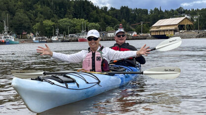 Two people in a tandem kayak paddle on calm water near a marina with boats and a boathouse visible in the background. They are smiling, and the front person has their arms outstretched, savoring the adventures in Canada’s scenic Bay of Fundy.