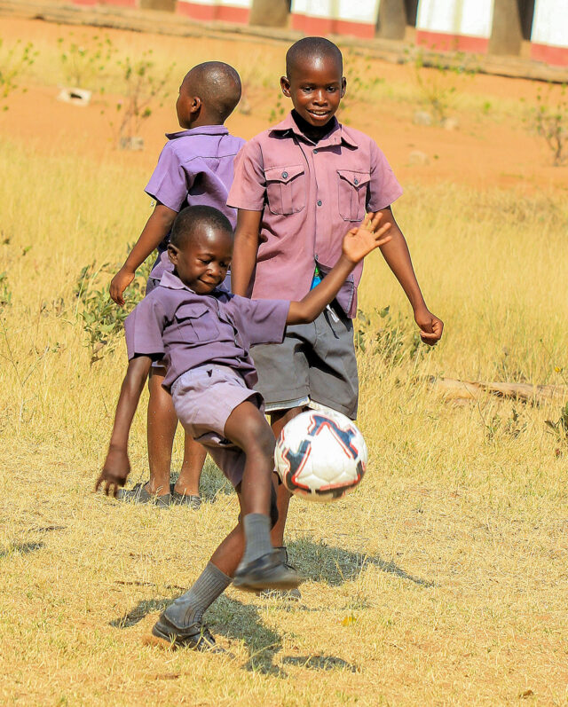 Three children in matching school uniforms are playing soccer outdoors on a sunny day, creating a scene that feels like heaven on Earth. One child is kicking a soccer ball while the other two watch and smile, their joy reminiscent of simpler times far from the remotest camp in Zimbabwe.