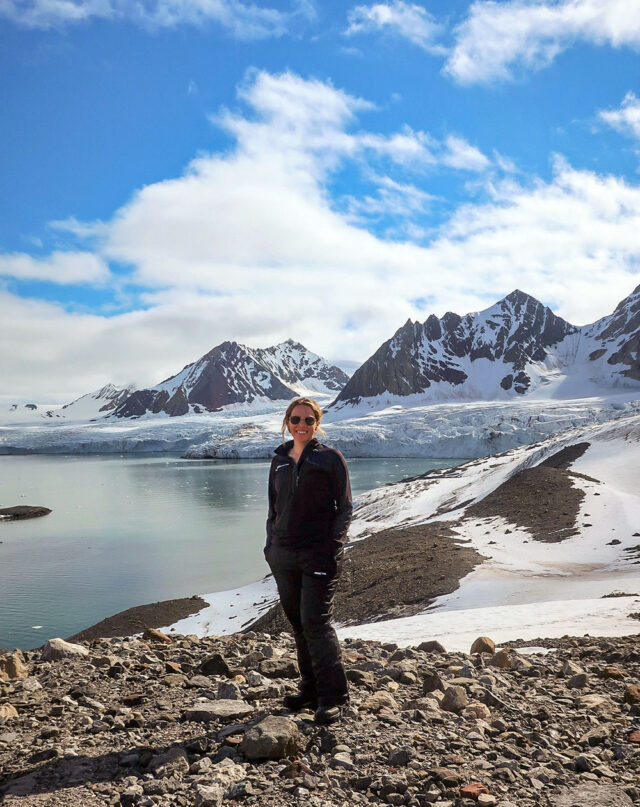 A person standing on rocky terrain with snowy mountains and a glacier in the background under a partly cloudy sky, capturing the essence of a Wildlife Adventure in Svalbard.