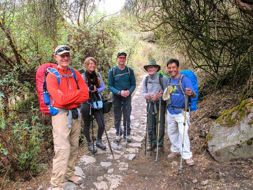 A group of five hikers, dressed in varying outdoor gear and carrying essential gear, stands on a forest path surrounded by greenery.
