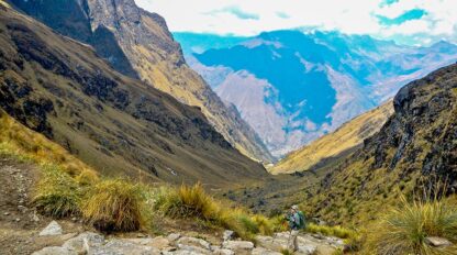 A hiker with a backpack, carrying all the hiking essentials, walks down the rocky Inca Trail, surrounded by steep cliffs and valleys under a partly cloudy sky.