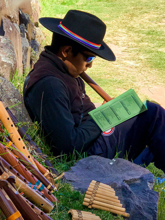 A person wearing a black hat with a colorful band sits outdoors, reading a book and holding a musical instrument after hiking the Inca Trail. Various wooden wind instruments are laid out beside them, reminiscent of the rich heritage found near Machu Picchu.