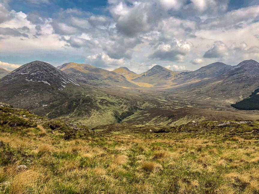 A scenic view of a mountainous landscape under a partly cloudy sky, featuring grassy slopes and rugged terrain reminiscent of Ireland's West Coast peaks.