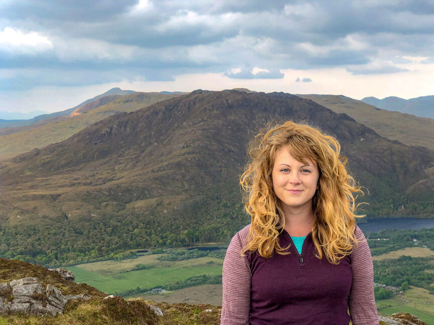 A woman with curly blonde hair stands in front of a scenic mountain landscape and cloudy sky. She is wearing a long-sleeved, half-zip top, perfectly suited for exploring Ireland's West Coast.