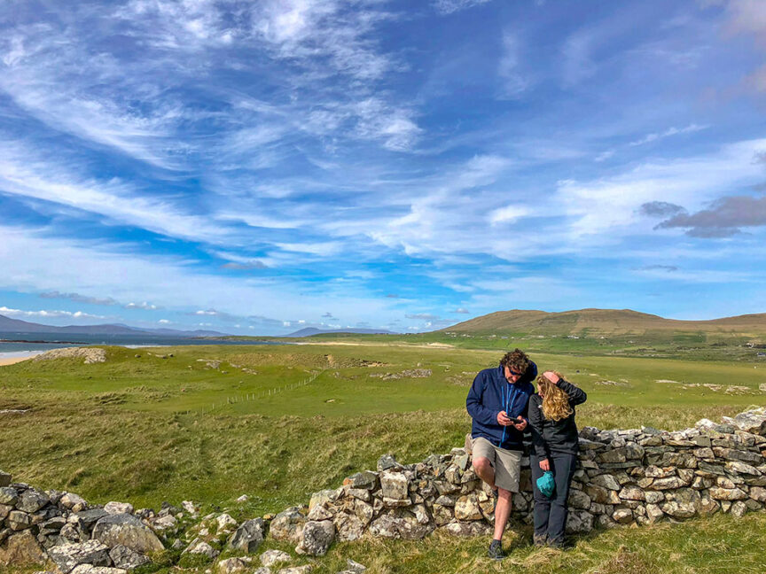 Two people leaning against a stone wall in a hilly, green landscape under a blue sky with scattered clouds on Ireland's West Coast. One person is looking at their phone while the other has an arm around them.