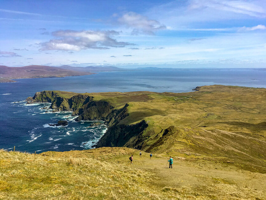 A group of hikers walks down a grassy hill towards Ireland's West Coast, where rugged cliffs and ocean waves meet a secluded beach under a partly cloudy sky.