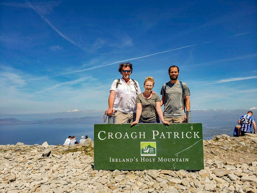 Three people stand behind a green sign reading "Croagh Patrick, Ireland's Holy Mountain." They are on a rocky summit with a clear sky and distant landscape in the background, epitomizing the essence of Exploring Ireland from Beach to Peaks.